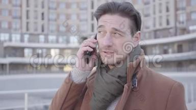 身着棕色外套的英俊自信男子站在城市街道上用手机交谈的肖像。 城市景观